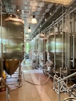 A modern brewery room with large stainless steel fermentation tanks lined up in a row. The room features a high ceiling with metal panels and industrial lighting. The floor is clean and equipped with drainage channels, and there are various pipes and hoses connected to the tanks.