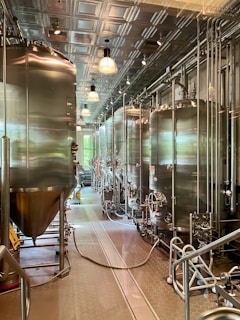 A modern brewery room with large stainless steel fermentation tanks lined up in a row. The room features a high ceiling with metal panels and industrial lighting. The floor is clean and equipped with drainage channels, and there are various pipes and hoses connected to the tanks.