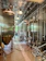 A modern brewery room with large stainless steel fermentation tanks lined up in a row. The room features a high ceiling with metal panels and industrial lighting. The floor is clean and equipped with drainage channels, and there are various pipes and hoses connected to the tanks.