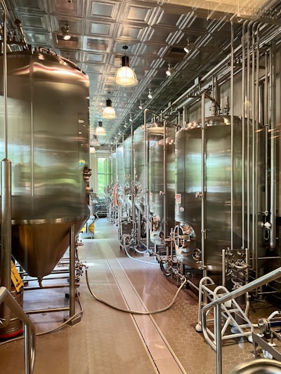 A modern brewery room with large stainless steel fermentation tanks lined up in a row. The room features a high ceiling with metal panels and industrial lighting. The floor is clean and equipped with drainage channels, and there are various pipes and hoses connected to the tanks.