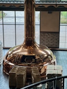 A large copper brewing kettle is situated in a room with high windows, allowing natural light to illuminate its polished surface. Numerous stacked bags, possibly containing ingredients, are arranged neatly in front of the kettle. The flooring consists of dark tiles, and the exterior view is partially visible through the windows.