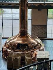A large copper brewing kettle is situated in a room with high windows, allowing natural light to illuminate its polished surface. Numerous stacked bags, possibly containing ingredients, are arranged neatly in front of the kettle. The flooring consists of dark tiles, and the exterior view is partially visible through the windows.