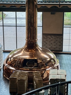 A large copper brewing kettle is situated in a room with high windows, allowing natural light to illuminate its polished surface. Numerous stacked bags, possibly containing ingredients, are arranged neatly in front of the kettle. The flooring consists of dark tiles, and the exterior view is partially visible through the windows.