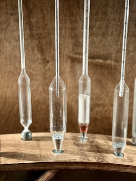 Several narrow glass tubes with varying amounts of granular material are mounted vertically on a wooden base. Each tube features measurement marks, suggesting a scientific or laboratory setting.