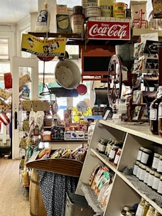 A nostalgic general store displaying a variety of vintage products and advertisements. Shelves are filled with jars of preserves and snacks, and old-fashioned signs such as Coca-Cola and Donald Duck Bread add a retro charm. The store has a cluttered but welcoming atmosphere, with barrels, bags of popcorn, and a colorful assortment of goods.