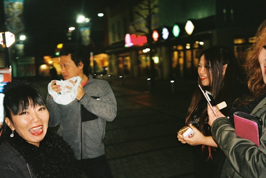 A group of people are gathered outdoors at night, with brightly lit signs in the background. One person is holding a napkin, while others are smiling and appear to be engaged in conversation.