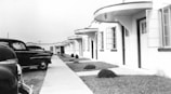 A vintage scene featuring a row of identical, single-story motel rooms with prominent rounded overhangs. The facade has several windows and doors, with small bushes neatly aligned in a grassy area adjacent to a paved walkway. On the left, several classic cars from the mid-20th century are parked, suggesting a retro setting.