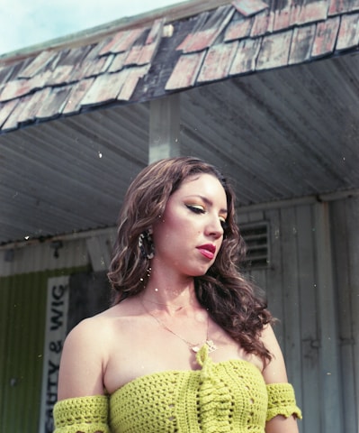 A woman with long, flowing hair is wearing a green crocheted top. She stands in front of an old, weathered building with a partially visible sign that reads 'Beauty & Wig'. The overhang of the building has a worn, paint-chipped appearance.