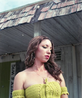 A woman with long, flowing hair is wearing a green crocheted top. She stands in front of an old, weathered building with a partially visible sign that reads 'Beauty & Wig'. The overhang of the building has a worn, paint-chipped appearance.