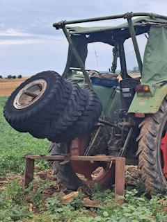 A green tractor positioned in a rural setting with its rear wheel elevated on a metal stand. The tractor tire has been removed and is placed nearby. The background features expansive farmland and a clear sky, with a wind turbine visible in the distance.