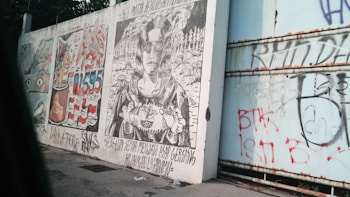 Graffiti and artwork cover a large outdoor wall. The wall features intricate black and white line art depicting a person wearing glasses with a detailed background of trees and possibly abstract elements. Next to this, there is colorful graffiti displaying numbers and abstract forms, including the appearance of flags or national symbols. The wall contains various tags, doodles, and text in different colors.