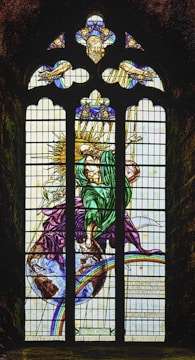 A stained glass window depicting a figure cloaked in green and purple, standing atop a representation of the Earth with a radiant sun behind. The person appears to be holding a staff or rod. Above, there are smaller sections showing hands and a face, set against a celestial theme with beams of light.