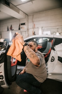 A man is cleaning the interior of a car door using an orange cloth. He appears to be in a garage or workshop setting, surrounded by various tools and equipment.