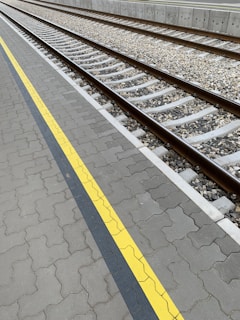 A railway track runs parallel to a textured platform with a yellow safety line. The track is made of wooden sleepers and gravel ballast, with metal rails. The platform features interlocking concrete pavers and a concrete barrier in the background.
