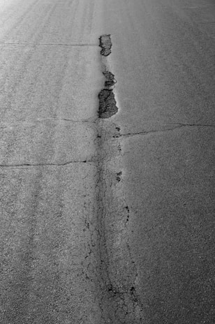 A cracked asphalt road with a prominent vertical fissure running through the center. The pavement surface displays various textures, with the crack showing signs of erosion and wear.