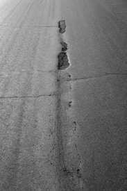 A cracked asphalt road with a prominent vertical fissure running through the center. The pavement surface displays various textures, with the crack showing signs of erosion and wear.