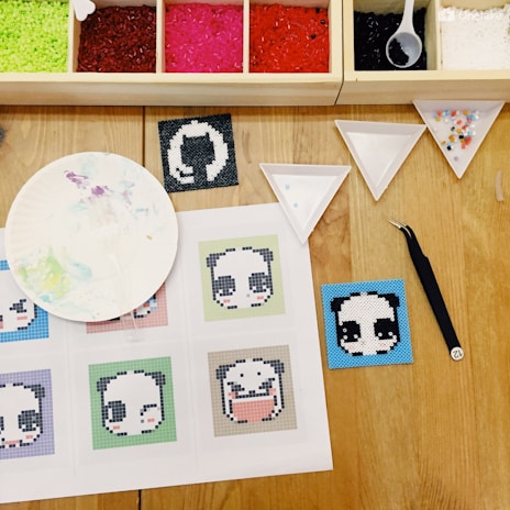 The image features a craft setup on a wooden table, including a variety of colored beads in a compartmentalized tray and tools such as tweezers and trays. Several panda-themed pixel art designs are displayed on a sheet of paper along with a palette plate with paint stains. A completed pixel art square with a panda face is seen next to the paper.