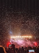 A large crowd of people is gathered in front of a stage with bright lights and colorful confetti filling the air. The audience holds up phones, capturing the moment, while the stage is illuminated with intense lighting, creating a vibrant and celebratory atmosphere.