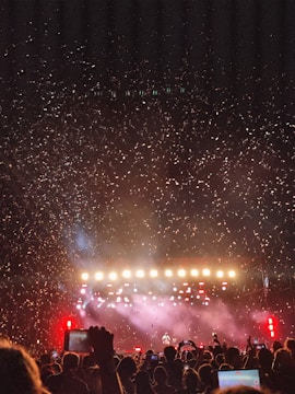 A large crowd of people is gathered in front of a stage with bright lights and colorful confetti filling the air. The audience holds up phones, capturing the moment, while the stage is illuminated with intense lighting, creating a vibrant and celebratory atmosphere.