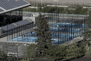 Aerial view of an outdoor sports complex featuring multiple enclosed courts with blue surfaces, surrounded by tall black fences. Several people are actively playing on the courts. Nearby is a large building with a sign reading 'MANIAK' and a tree in the foreground. Solar panels are visible on the roof of the structure beside the courts.