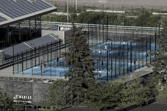 Aerial view of an outdoor sports complex featuring multiple enclosed courts with blue surfaces, surrounded by tall black fences. Several people are actively playing on the courts. Nearby is a large building with a sign reading 'MANIAK' and a tree in the foreground. Solar panels are visible on the roof of the structure beside the courts.