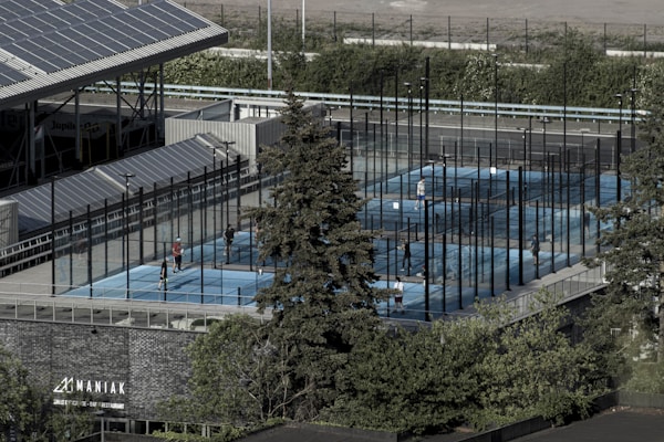 Aerial view of an outdoor sports complex featuring multiple enclosed courts with blue surfaces, surrounded by tall black fences. Several people are actively playing on the courts. Nearby is a large building with a sign reading 'MANIAK' and a tree in the foreground. Solar panels are visible on the roof of the structure beside the courts.