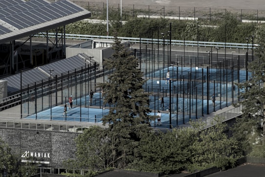 Aerial view of an outdoor sports complex featuring multiple enclosed courts with blue surfaces, surrounded by tall black fences. Several people are actively playing on the courts. Nearby is a large building with a sign reading 'MANIAK' and a tree in the foreground. Solar panels are visible on the roof of the structure beside the courts.