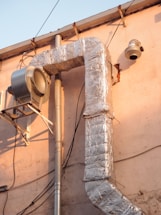 A large, metallic duct system is attached to an exterior wall of a building. The ductwork is insulated with shiny, reflective silver material and connected to a vent or fan unit. Various electrical wires are visible running alongside the structure, and the building's wall has a light pink hue, bathed in warm sunlight.