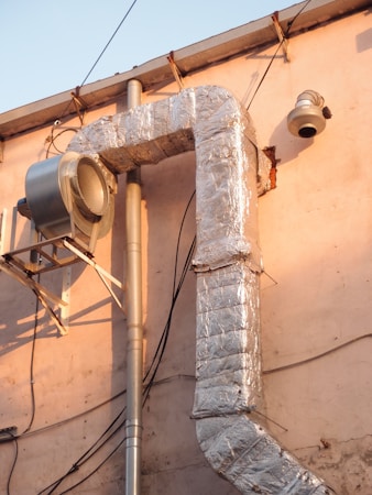 A large, metallic duct system is attached to an exterior wall of a building. The ductwork is insulated with shiny, reflective silver material and connected to a vent or fan unit. Various electrical wires are visible running alongside the structure, and the building's wall has a light pink hue, bathed in warm sunlight.