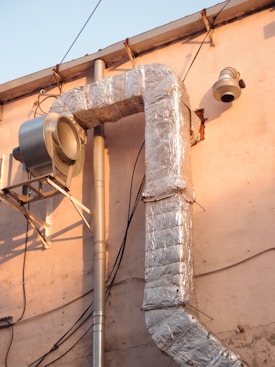 A large, metallic duct system is attached to an exterior wall of a building. The ductwork is insulated with shiny, reflective silver material and connected to a vent or fan unit. Various electrical wires are visible running alongside the structure, and the building's wall has a light pink hue, bathed in warm sunlight.