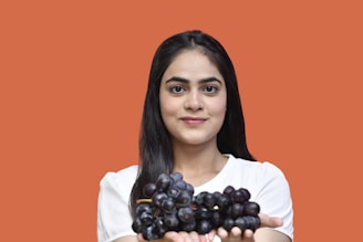 A woman with long dark hair is holding a bunch of black grapes in her hands. She is wearing a white shirt and is set against a solid orange background. Her expression is calm and serene, with a gentle smile.