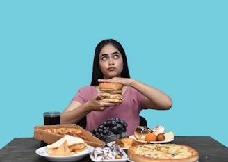 A person with long dark hair holds a large hamburger with both hands, looking upwards in a contemplative expression. The table in front displays a variety of foods including pizza, a sandwich, grapes, assorted pastries, and a drink. The background is a solid teal color.