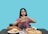 A person with long dark hair holds a large hamburger with both hands, looking upwards in a contemplative expression. The table in front displays a variety of foods including pizza, a sandwich, grapes, assorted pastries, and a drink. The background is a solid teal color.