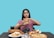 A person with long dark hair holds a large hamburger with both hands, looking upwards in a contemplative expression. The table in front displays a variety of foods including pizza, a sandwich, grapes, assorted pastries, and a drink. The background is a solid teal color.
