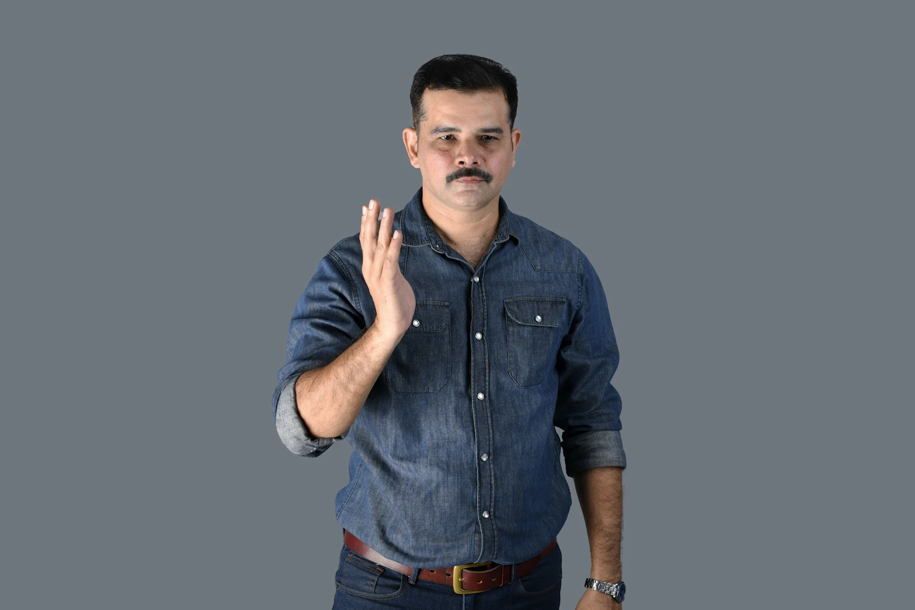 A man in a denim shirt holding one open hand up beside his face, in front of a plain gray backdrop