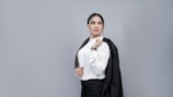 A person with long dark hair is dressed in a white button-up shirt and holding a black blazer over one shoulder. They have a confident expression and are standing against a plain gray background.