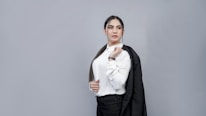 A person with long dark hair is dressed in a white button-up shirt and holding a black blazer over one shoulder. They have a confident expression and are standing against a plain gray background.