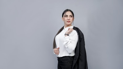 A person with long dark hair is dressed in a white button-up shirt and holding a black blazer over one shoulder. They have a confident expression and are standing against a plain gray background.