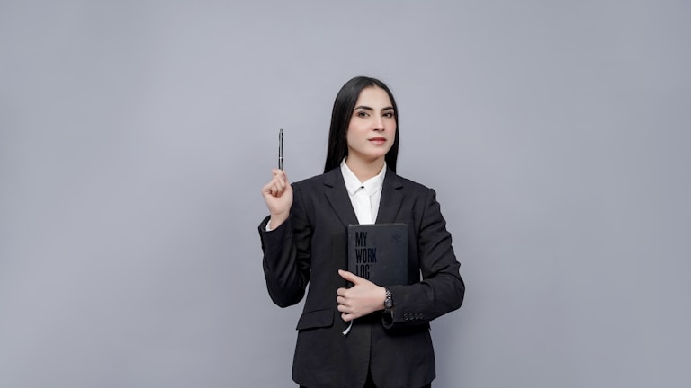 A person dressed in a formal suit holding a notebook labeled 'My Work Log' against their chest, while also holding a pen in their raised hand. The background is a plain gray color, creating a professional and minimalist look.