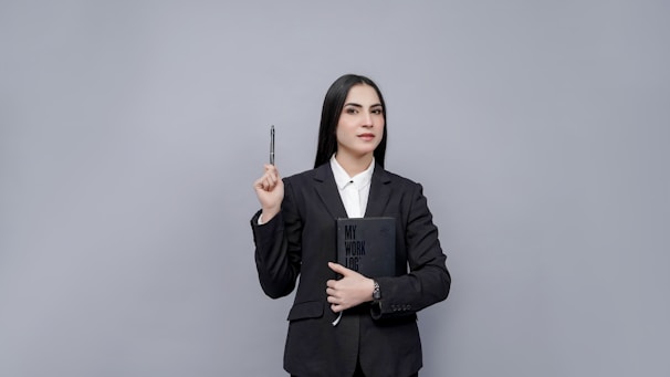 A person dressed in a formal suit holding a notebook labeled 'My Work Log' against their chest, while also holding a pen in their raised hand. The background is a plain gray color, creating a professional and minimalist look.