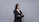 A professional woman in a black suit and white shirt stands with arms crossed against a plain gray background. Her long dark hair is straight and she exudes confidence.