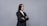 A professional woman in a black suit and white shirt stands with arms crossed against a plain gray background. Her long dark hair is straight and she exudes confidence.