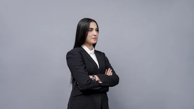A confident woman in a tailored blazer and trousers standing against a minimalist backdrop.
