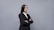 A professional woman in a black suit and white shirt stands with arms crossed against a plain gray background. Her long dark hair is straight and she exudes confidence.