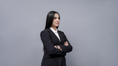 A professional woman in a black suit and white shirt stands with arms crossed against a plain gray background. Her long dark hair is straight and she exudes confidence.