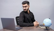 A man with glasses sits at a desk, looking to the side. He wears a black shirt and appears to be thoughtful. In front of him is a laptop, a globe, a book, and a pen on the wooden table. The background is a plain gray color.