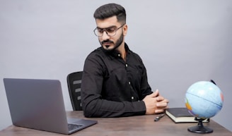 A thoughtful older man writing notes at a desk surrounded by books and a laptop.