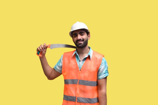 A person wearing an orange reflective safety vest and a white hard hat is holding a handsaw with a curved blade. The background is solid yellow.