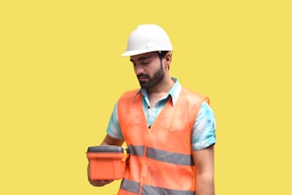 A male construction worker wearing a white hard hat and orange safety vest is holding an orange case. He is looking at the case, standing against a plain yellow background. The worker is also wearing a blue shirt under the vest.