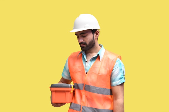 A male construction worker wearing a white hard hat and orange safety vest is holding an orange case. He is looking at the case, standing against a plain yellow background. The worker is also wearing a blue shirt under the vest.